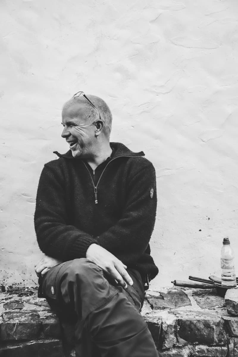 Dirk Ottenheym, black-and-white portrait, laughing, arms resting on his knees, against a white stucco wall.